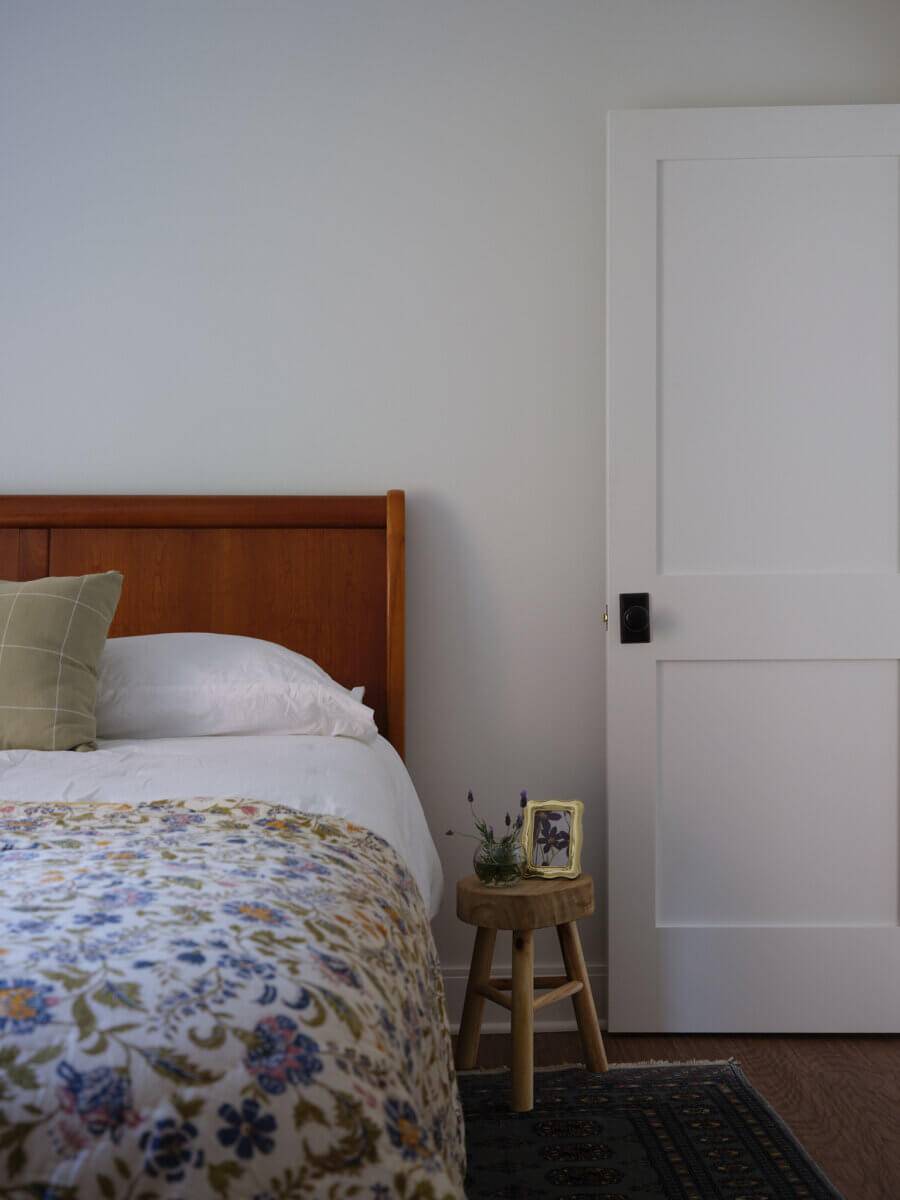 A neatly made bed with a wooden headboard sits next to a small stool holding a framed photo and a small plant, beside a white door in a simple bedroom.