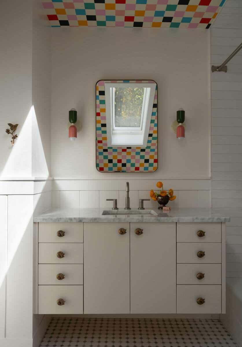A bathroom features a white vanity, marble countertop, geometric floor tiles, and a mirror framed by colorful checkerboard tiles that extend onto the ceiling. A skylight above the mirror brightens the space.
