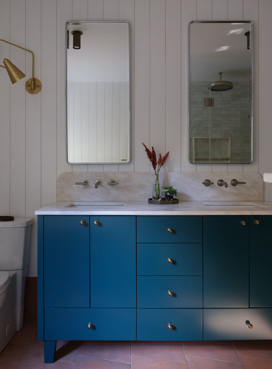 A modern bathroom features a blue double-sink vanity with gold hardware, two rectangular mirrors, a wall sconce, and terracotta floor tiles.