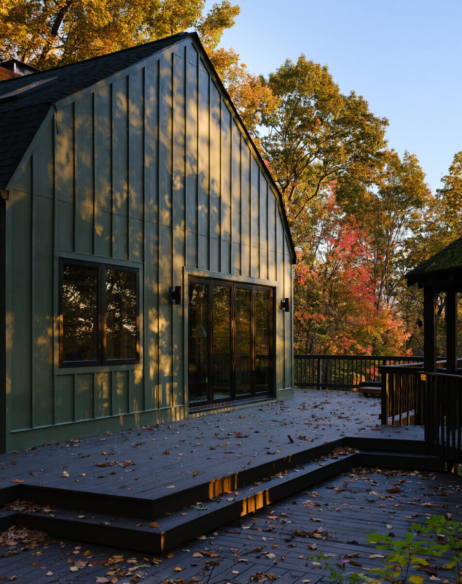 Modern house with vertical green siding, featuring large windows and a spacious deck covered with fallen autumn leaves, surrounded by trees in vibrant fall foliage.