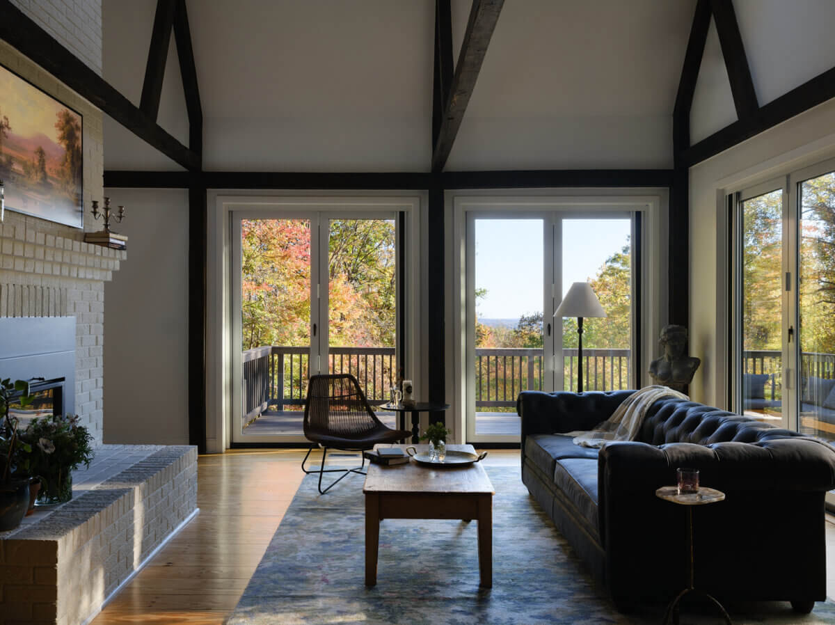 A cozy living room in the Marshall Barn features a dark sofa, wooden coffee table, armchair, and large windows revealing a deck and autumn trees outside. Sunlight streams through the glass doors.