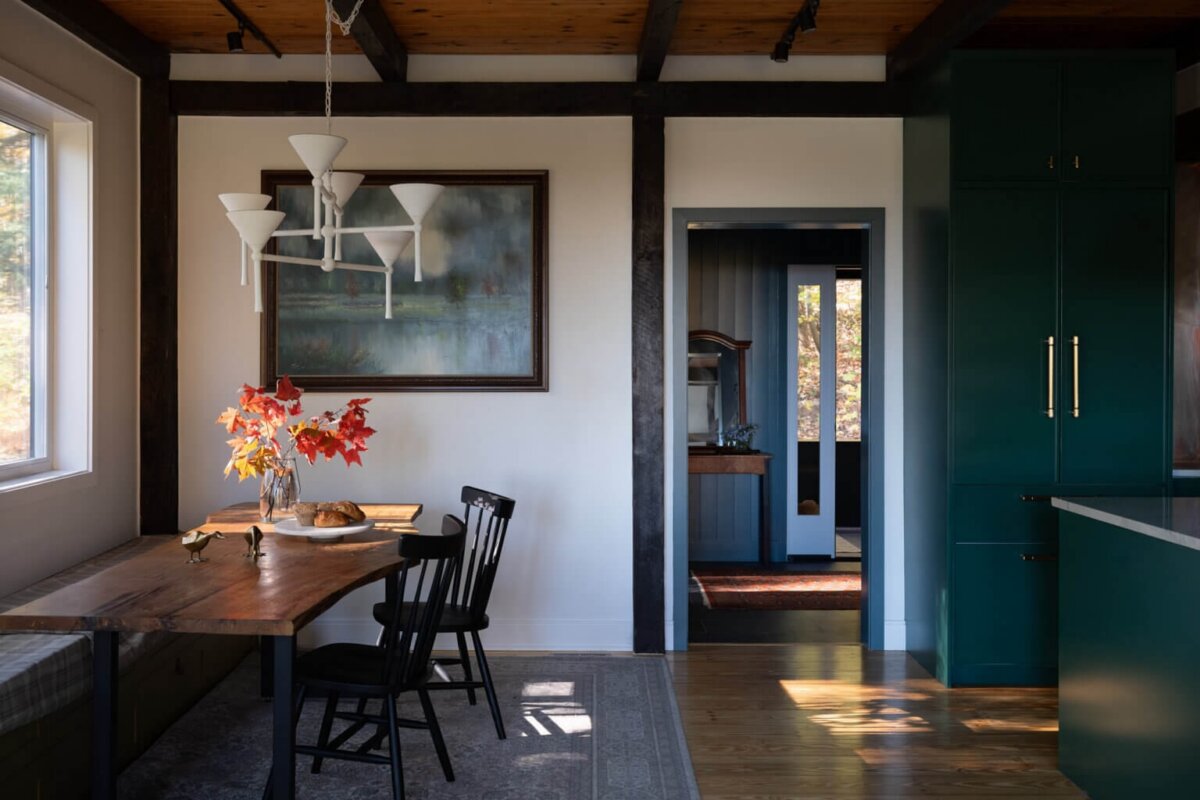 A dining area in a cozy barn home features a wooden table, black chairs, a vase of autumn leaves, green cabinets, and a large window letting in natural light.