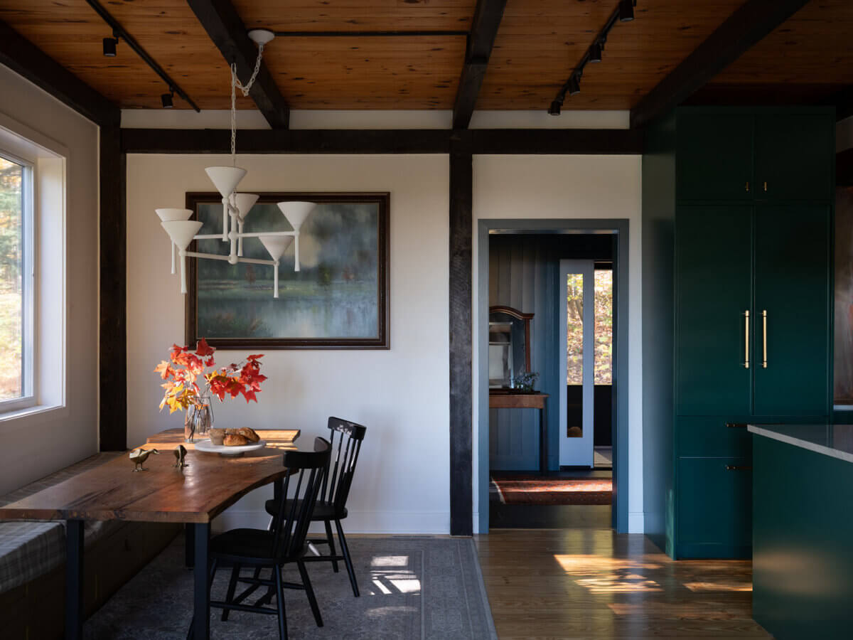 A dining area in Marshall Barn features a wooden table, black chairs, a vase of red leaves, chandelier, large painting, and green kitchen cabinets under a wooden beam ceiling.