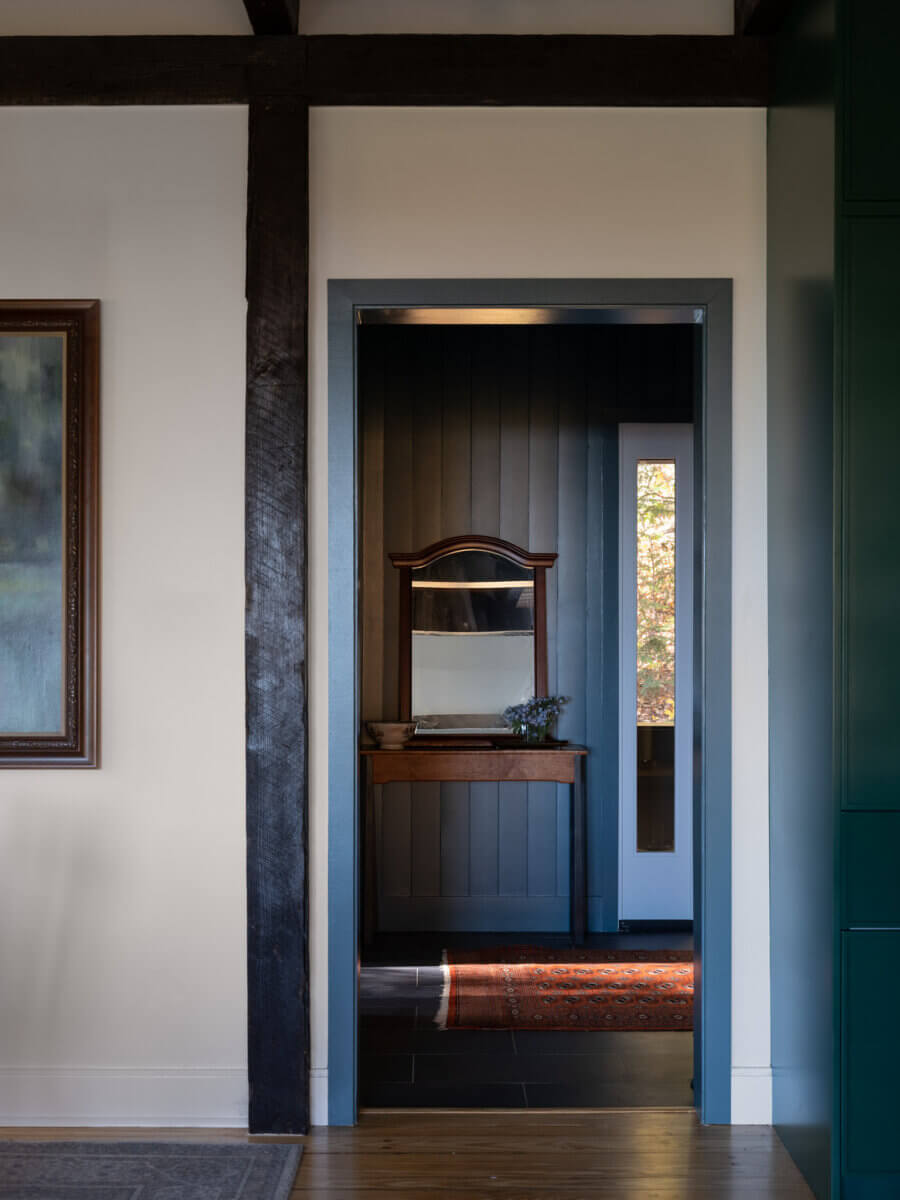 A wooden vanity with a mirror, small plant, and drawer sits in the dark-paneled room of The Marshall Barn, viewed through an open doorway from a well-lit adjacent space.