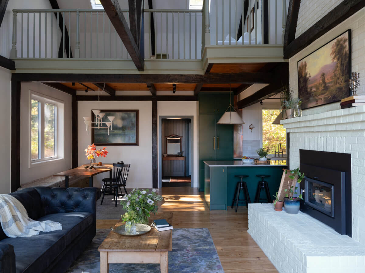 A modern living room at Marshall Barn features a black sofa, fireplace, coffee table with plants, open kitchen with green cabinets, dining area, and exposed wooden beams with a loft above.