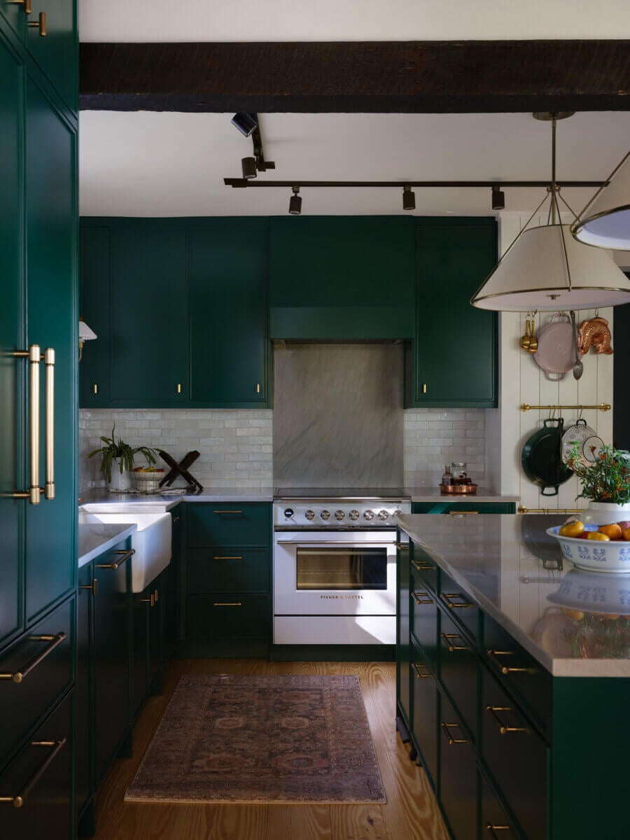 A modern kitchen in Marshall Barn featuring dark green cabinets, brass hardware, a stainless steel stove, white tile backsplash, farmhouse sink, pendant lights, and a rug on a wooden floor.