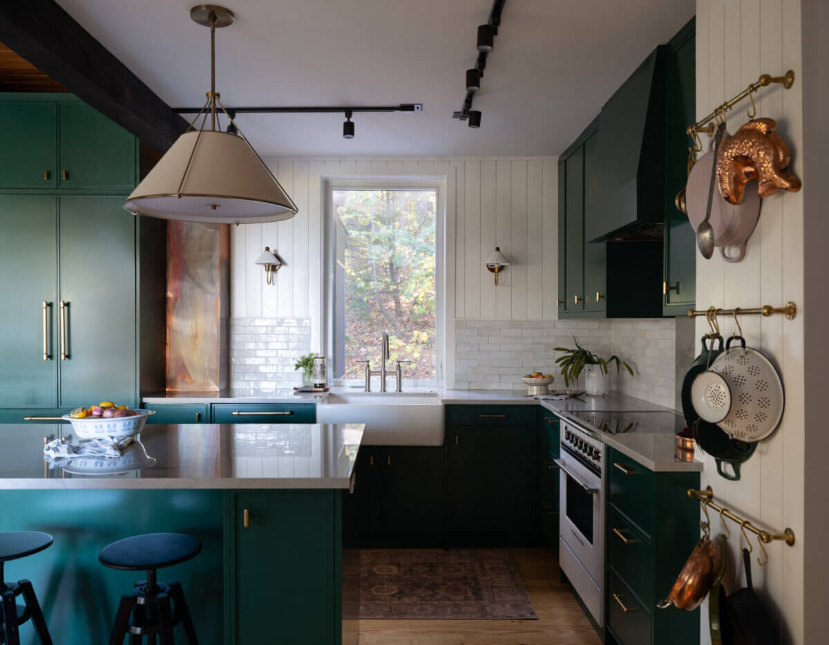 Modern kitchen with green cabinets, white countertops, and a central island with stools. Hanging pots and a large window overlooking trees fill the space with natural light.