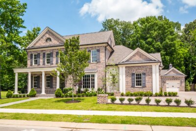 Two-story brick house by Magnolia Homes with a covered front porch, manicured lawn, shrubs, and a detached garage—an ideal setting for building lasting legacies beneath a partly cloudy sky and tall trees.
