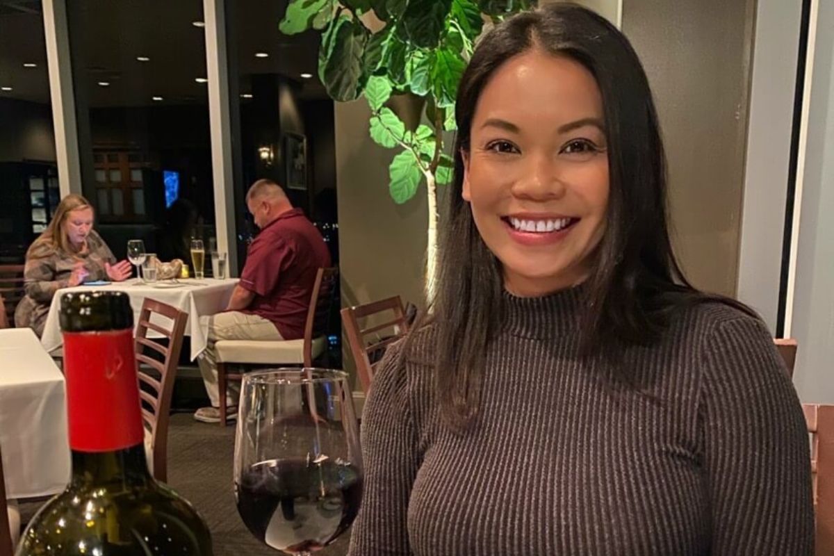 A woman sits at a Wine Not Lounge table with a glass and bottle of red wine, smiling at the camera as she celebrates her career shift; other diners are visible in the background.