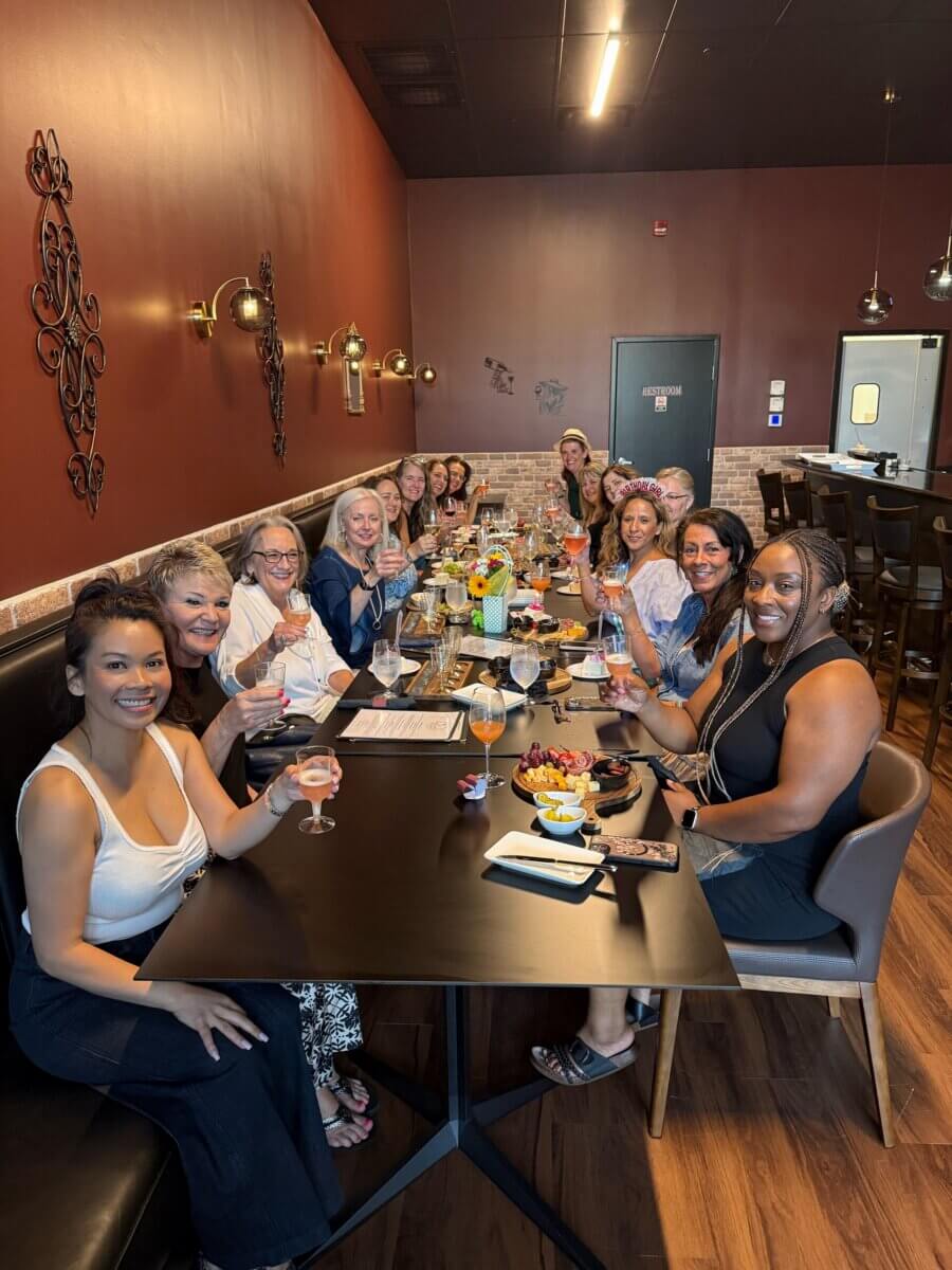 A group of women sit around a long table in a restaurant, smiling and raising glasses, with food and drinks in front of them.