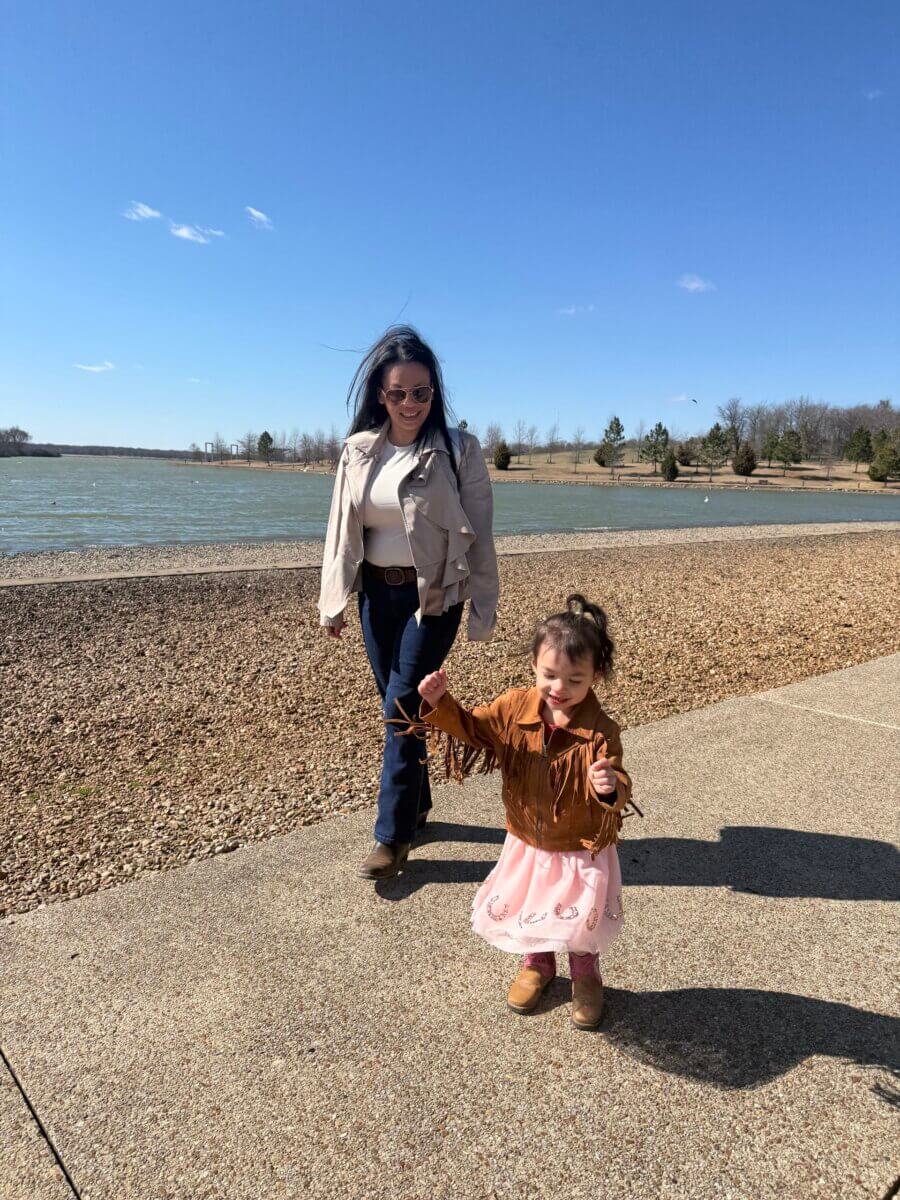 A woman and a young girl walk along a lakeside path on a sunny day, with the girl smiling and holding out her arms.
