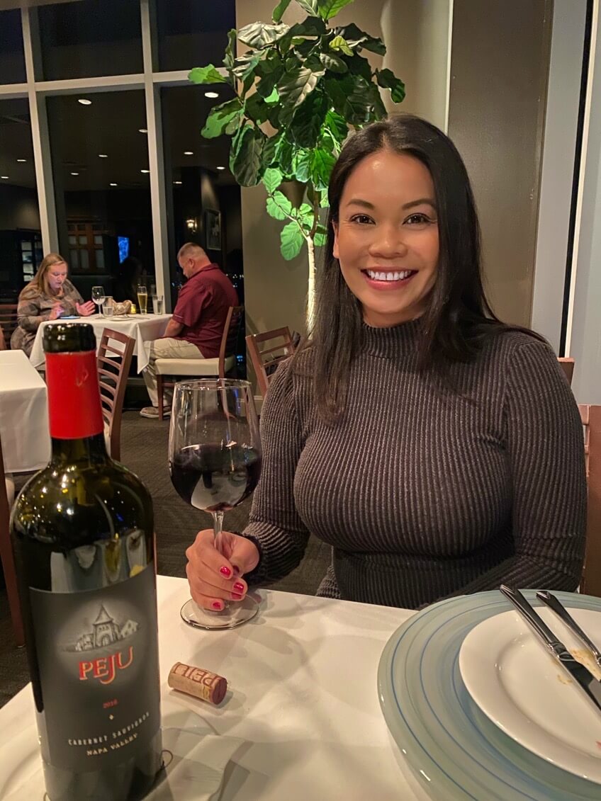 A woman sits at a restaurant table with a glass of red wine, a bottle of Peju wine, a cork, and place settings in front of her. She is smiling at the camera.