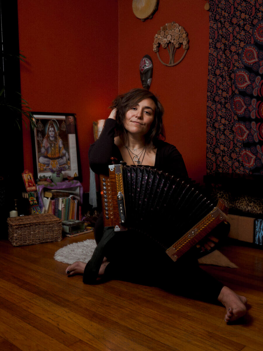A woman sits barefoot on a wooden floor in a cozy, decorated room, holding an accordion and smiling at the camera. Books, art, and musical instruments are visible in the background.