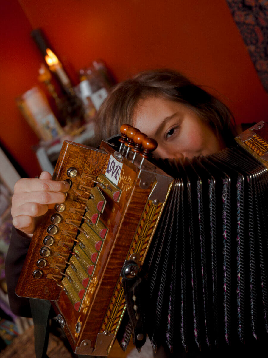 A person holding and playing a wooden accordion with brass buttons in a warmly lit room with an orange wall in the background.