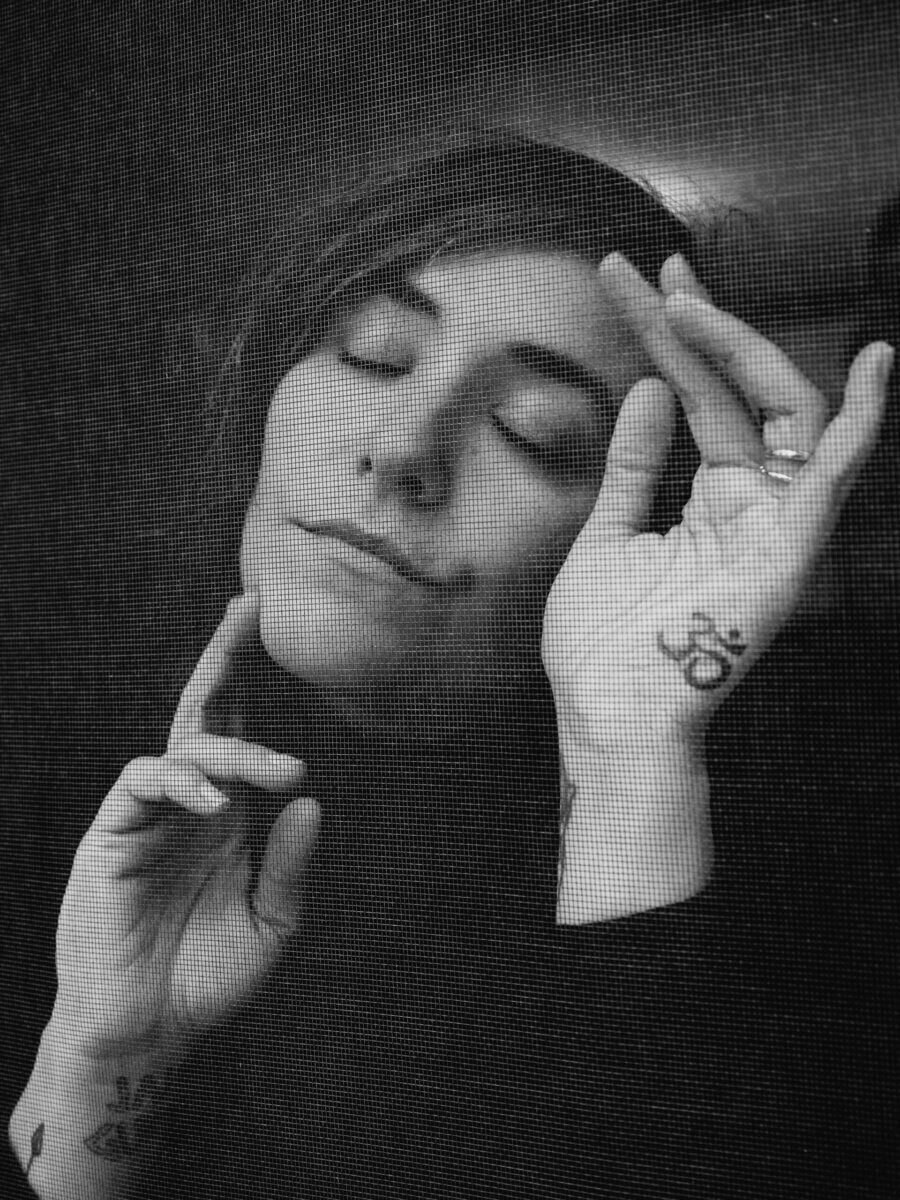 Woman with eyes closed poses behind a mesh screen, one hand raised near her face displaying a visible tattoo on her wrist.