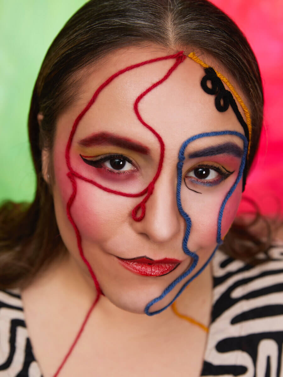 A woman with bold makeup and red, blue, and yellow yarn arranged in abstract patterns on her face, set against a blurred green and red background.