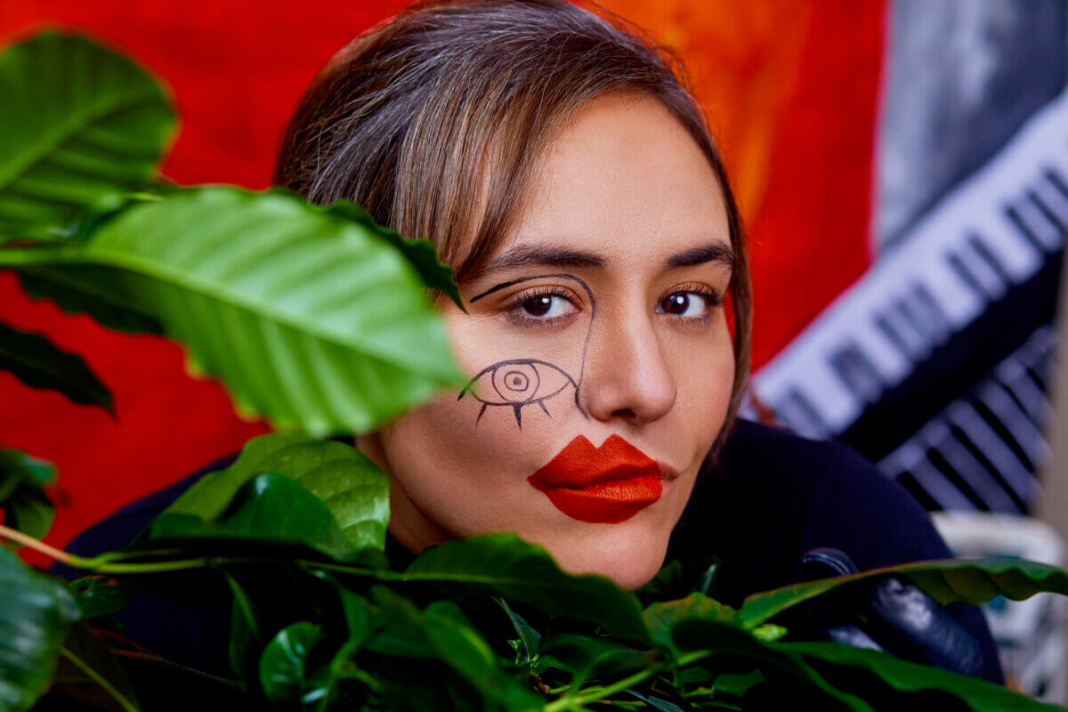 A woman with red lipstick and an eye drawn on her cheek looks at the camera, partially obscured by green leaves, against a vibrant background.