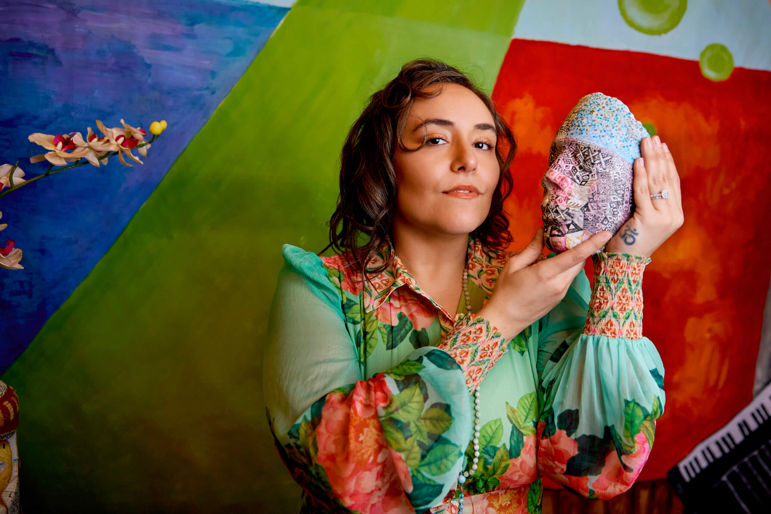A woman in a floral dress holds a decorated head sculpture against a colorful, geometric background.