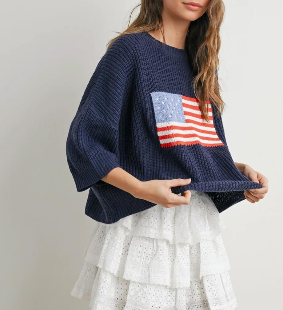 15 Memphis FINDS to Kick Off Summer: Vacation-Ready Style & More - 7 A woman wearing a navy blue knit sweater with an American flag design and a white tiered lace skirt stands against a plain background—perfect inspiration for Mother’s Day gift ideas.
