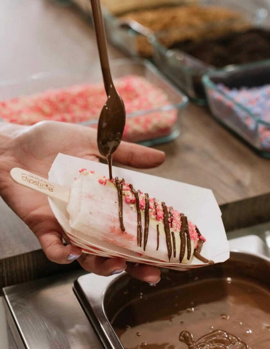 A hand holds a popsicle covered with pink sprinkles while chocolate sauce is being drizzled on top using a spoon, with topping containers in the background.