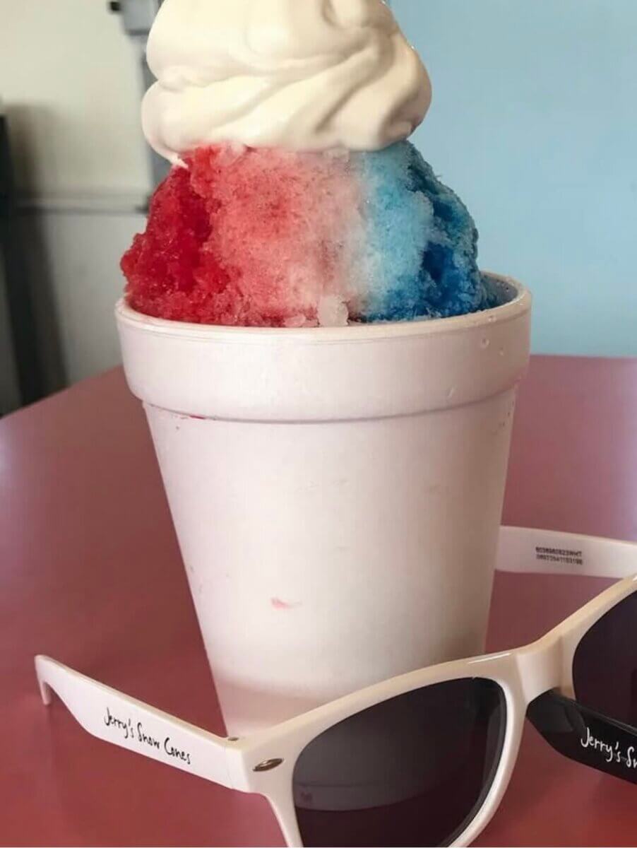 A red, white, and blue snow cone with soft serve ice cream on top in a white Styrofoam cup sits on a pink table next to white sunglasses.
