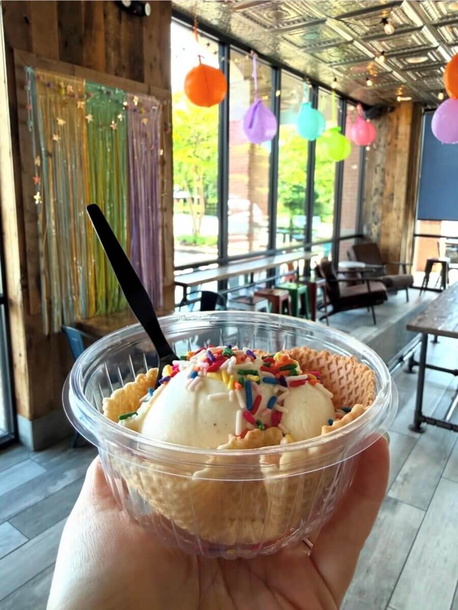 A hand holds a plastic bowl of vanilla ice cream with rainbow sprinkles and a waffle bowl, inside a decorated cafe with colorful balloons and streamers.