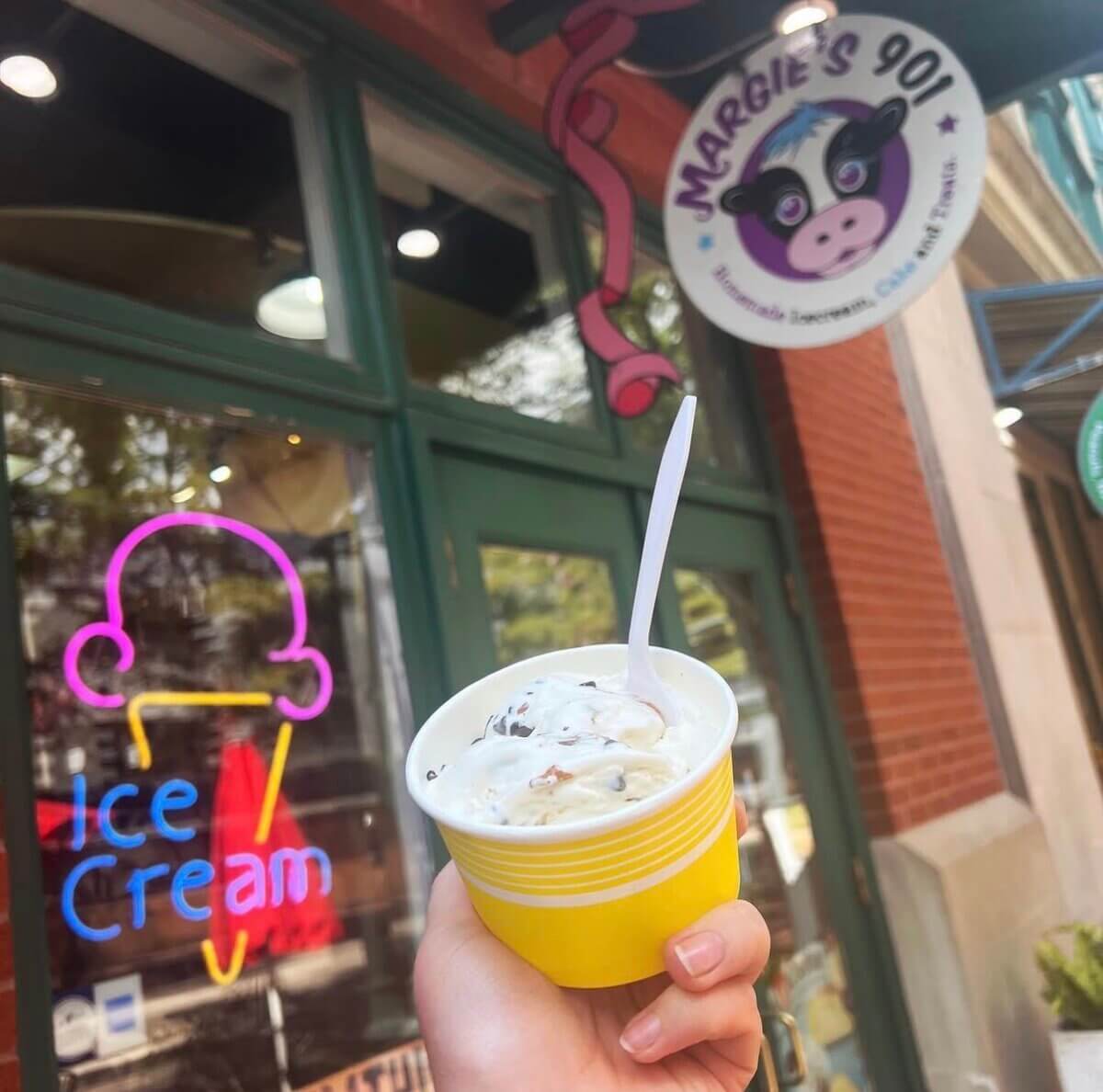 A hand holds a yellow cup of ice cream with a spoon in front of Margie’s 901 ice cream shop, with a neon ice cream sign visible on the window.