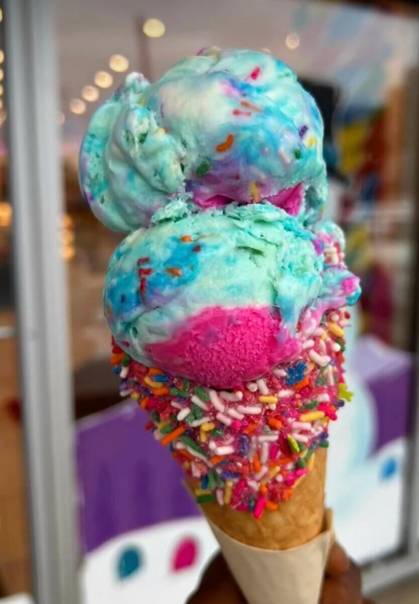 A waffle cone with two scoops of colorful ice cream, mostly blue and pink, covered in rainbow sprinkles.