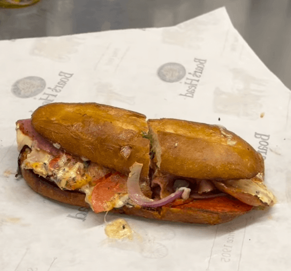 A toasted sandwich with meat, cheese, onions, and vegetables sits on branded deli paper on a counter.