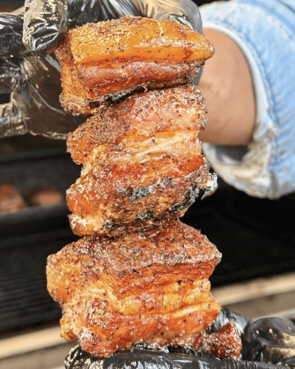 A gloved hand holds up three pieces of seasoned, grilled pork belly stacked on top of each other, with a smoker visible in the background.