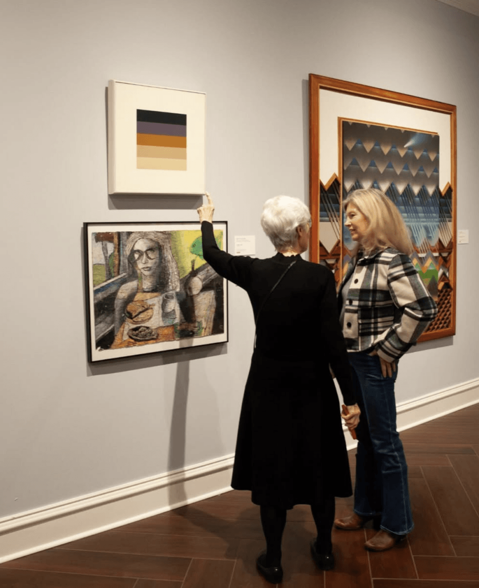 The South’s Biggest Museum Boom is Happening in Memphis - 2 Two women stand in an art gallery; one gestures toward an abstract painting while the other looks on. Several artworks hang on the wall behind them.
