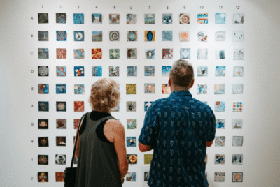 Two people stand in front of a wall displaying a grid of small, varied square artworks arranged in rows and columns.