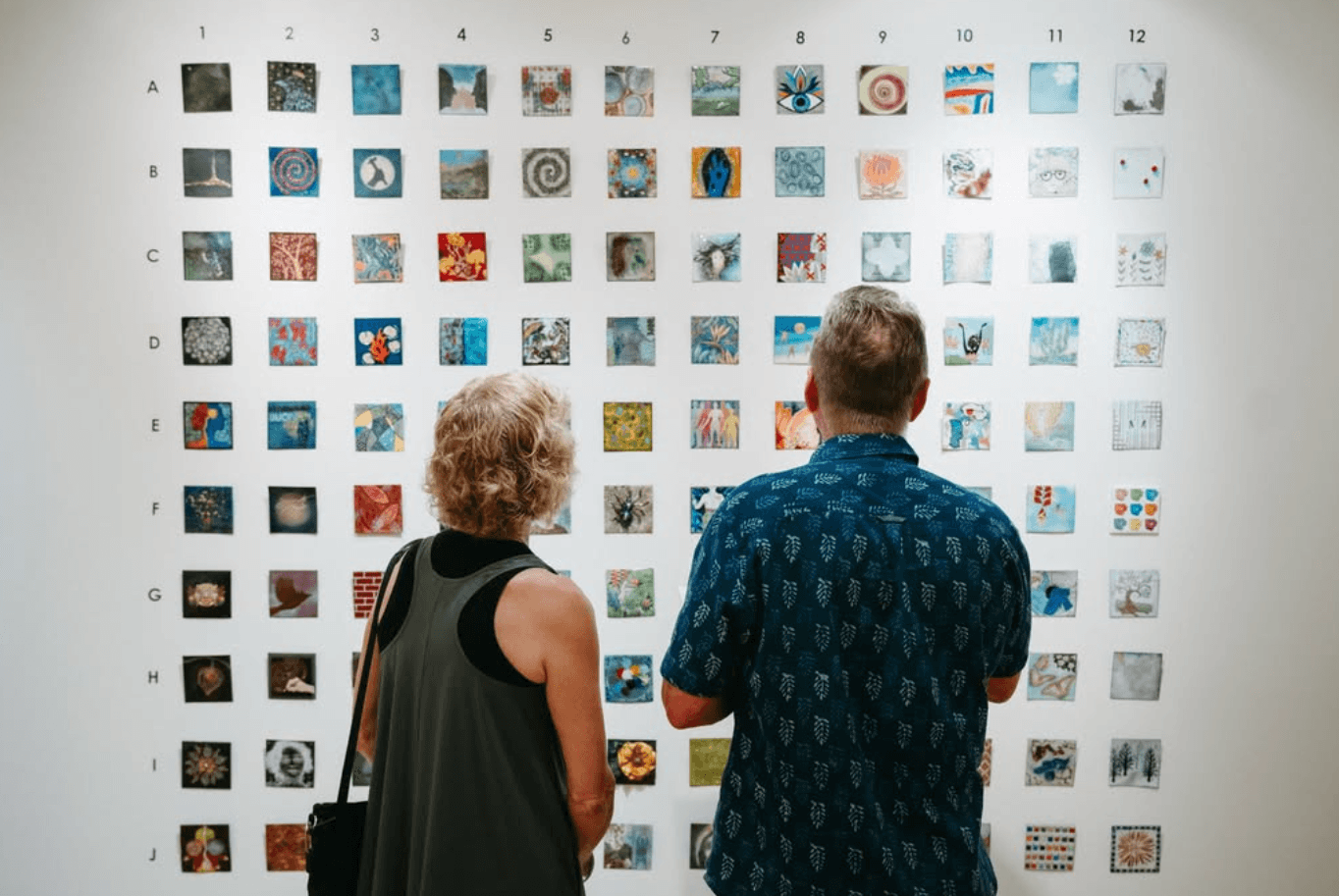 Two people stand in front of a wall displaying a grid of small, varied square artworks arranged in rows and columns.
