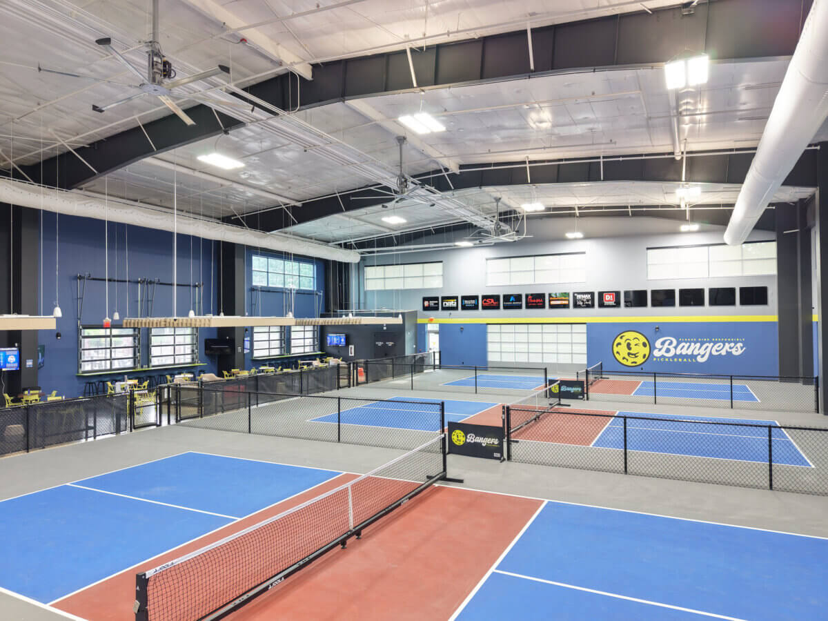 11 Places to Play Pickleball in Memphis - 3 Indoor pickleball facility in Memphis featuring multiple blue and red courts, high ceilings, scoreboard screens, and seating areas along the sides. A bold "Bangers" sign is displayed on the far wall.