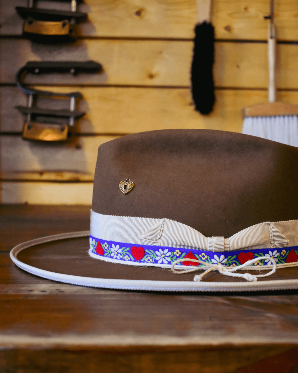 18 Nashville Luxe Loves: Statement Jewelry, Custom Hats & Summer Showstoppers - 5 A Luxe brown wide-brimmed hat with a white ribbon and colorful heart and flower trim sits on a wooden surface; hat-making tools hang on a wooden wall in the background, perfect for anyone who Loves Nashville style.