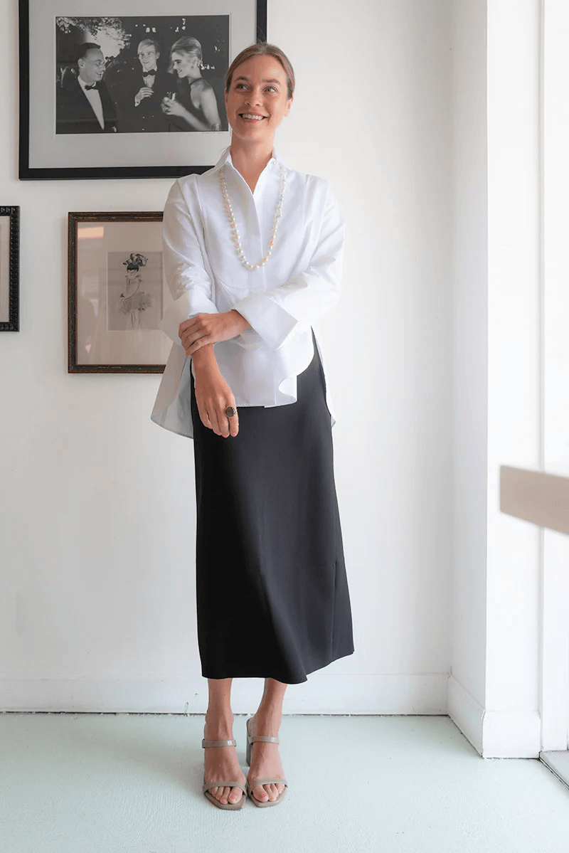 18 Nashville Luxe Loves: Statement Jewelry, Custom Hats & Summer Showstoppers - 3 A woman stands indoors in a luxe white blouse, black midi skirt, pearl necklace, and sandals. Framed photos decorate the white wall behind her.