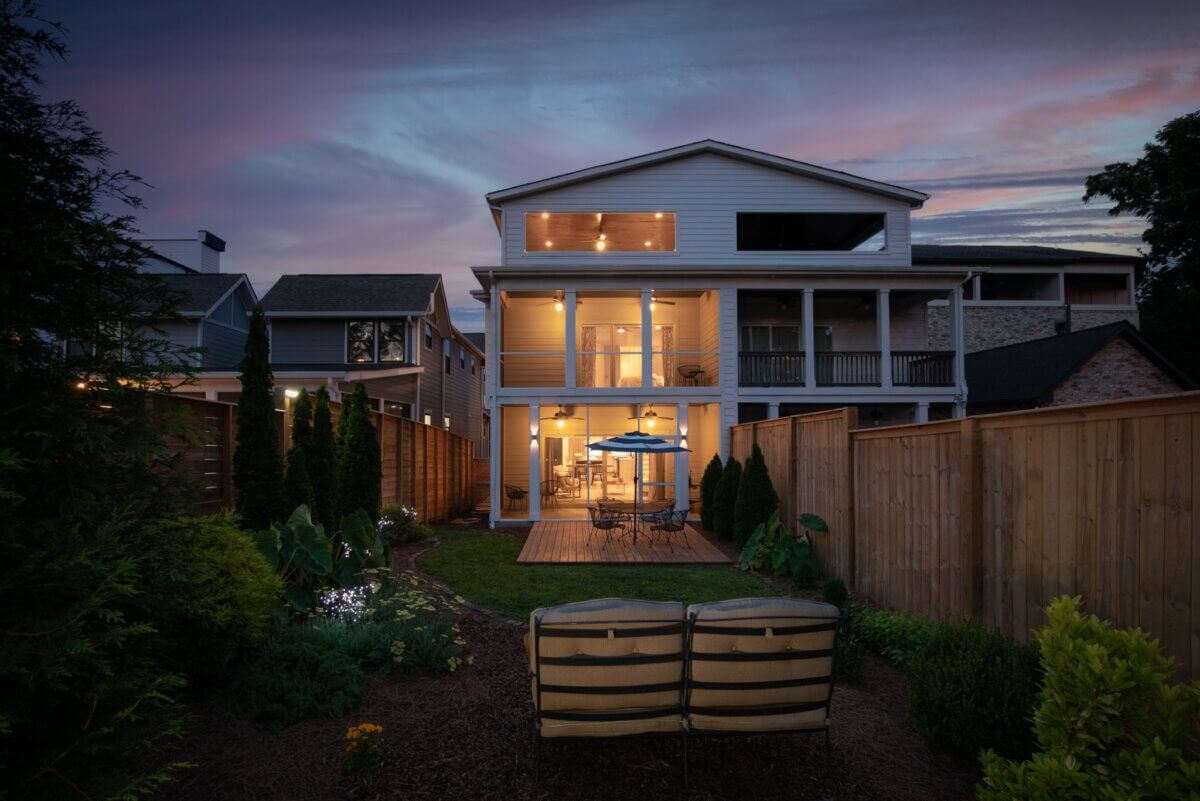 Backyard view of a modern two-story house at dusk on W Kirkwood Ave, Nashville—illuminated windows, a patio dining area under an umbrella, and two cushioned chairs facing the house create an inviting scene at 2815A.