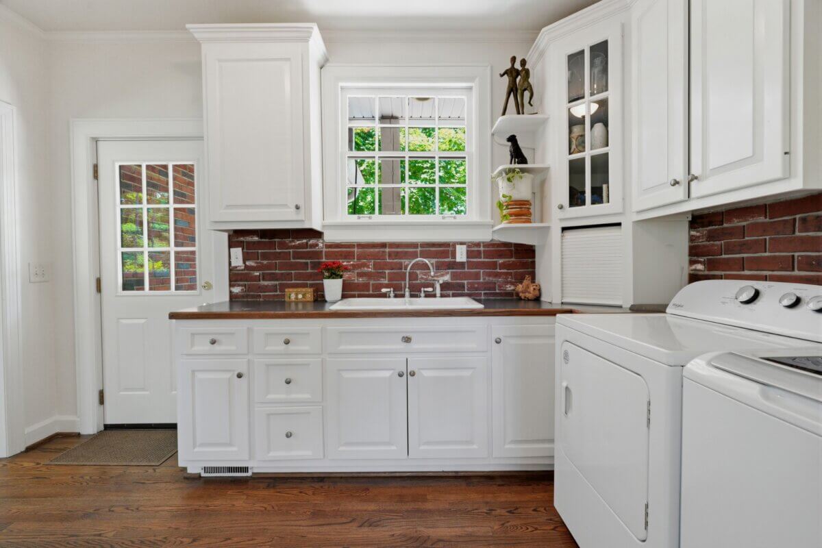 A bright kitchen with white cabinets, brick backsplash, wood countertop, a window above the sink, and a white washer and dryer next to the counter at 3008 Natchez Trce in Nashville TN.