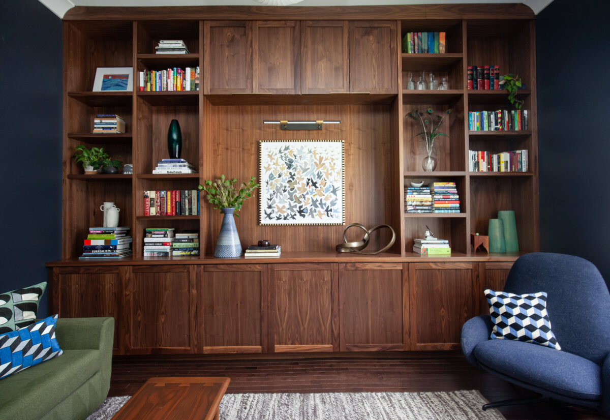 Built-in wooden bookshelves filled with books, plants, and decor items, centered by framed floral artwork above cabinets; a dark blue armchair and green sofa in front create a cozy space—perfect for considering when to hire an interior designer.