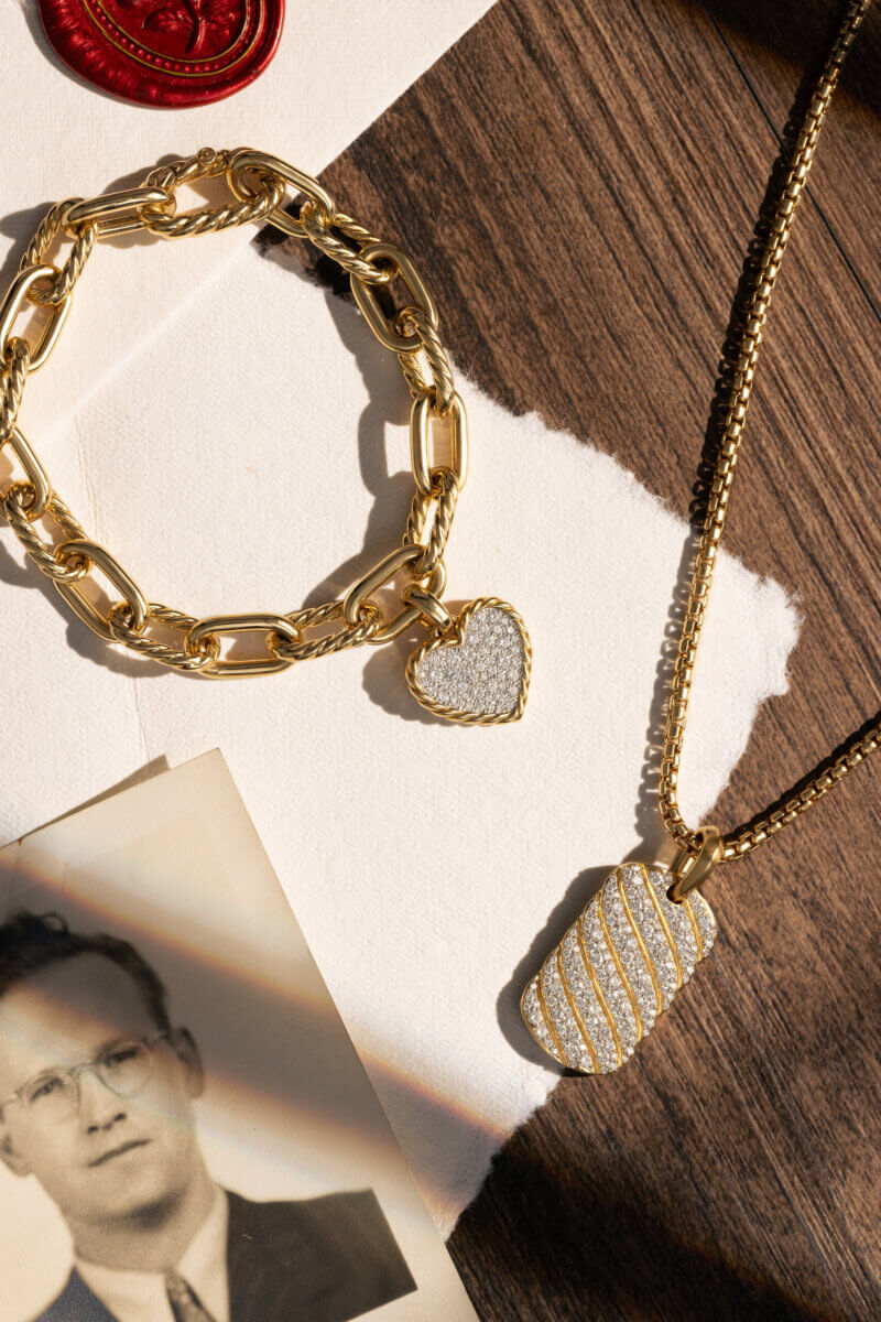 Personalized Jewelry Gifts for Life’s Biggest Milestones - 2 Gold chain bracelet with a heart-shaped charm and a gold necklace with a rectangular pendant on a wooden surface, next to a black-and-white portrait and torn paper.