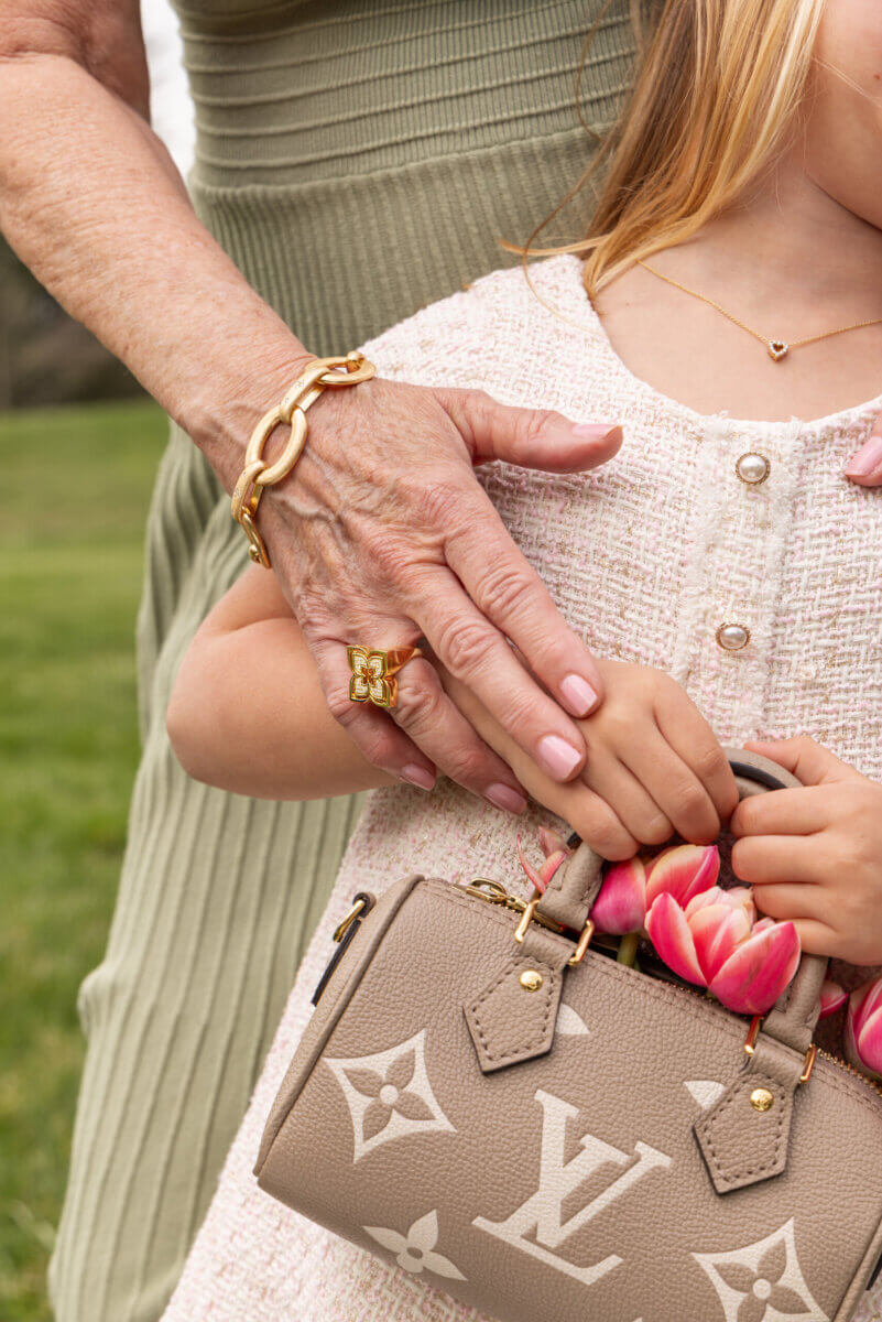 Personalized Jewelry Gifts for Life’s Biggest Milestones - 4 An older woman gently holds a young girl’s hands as she clutches a small Louis Vuitton handbag with pink tulips inside. Both wear gold and fine jewelry.