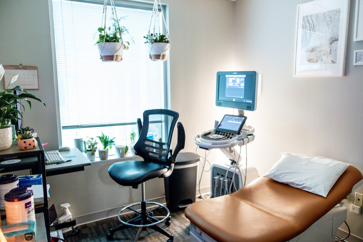 Beyond Birth: Why Women Are Choosing Midwives for Everyday Care - 3 A medical examination room designed for everyday care, featuring a brown exam table, black office chair, ultrasound machine, desk with computer, potted plants, and natural light from a window—ideal for women's healthcare visits with midwives.