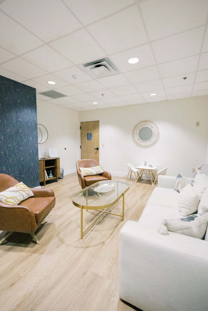 Beyond Birth: Why Women Are Choosing Midwives for Everyday Care - 6 A modern waiting room designed for everyday care, featuring a white sofa, two brown chairs, a glass coffee table, and light wood flooring; wall art and mirrors add warmth to this inviting space often visited by women and midwives.