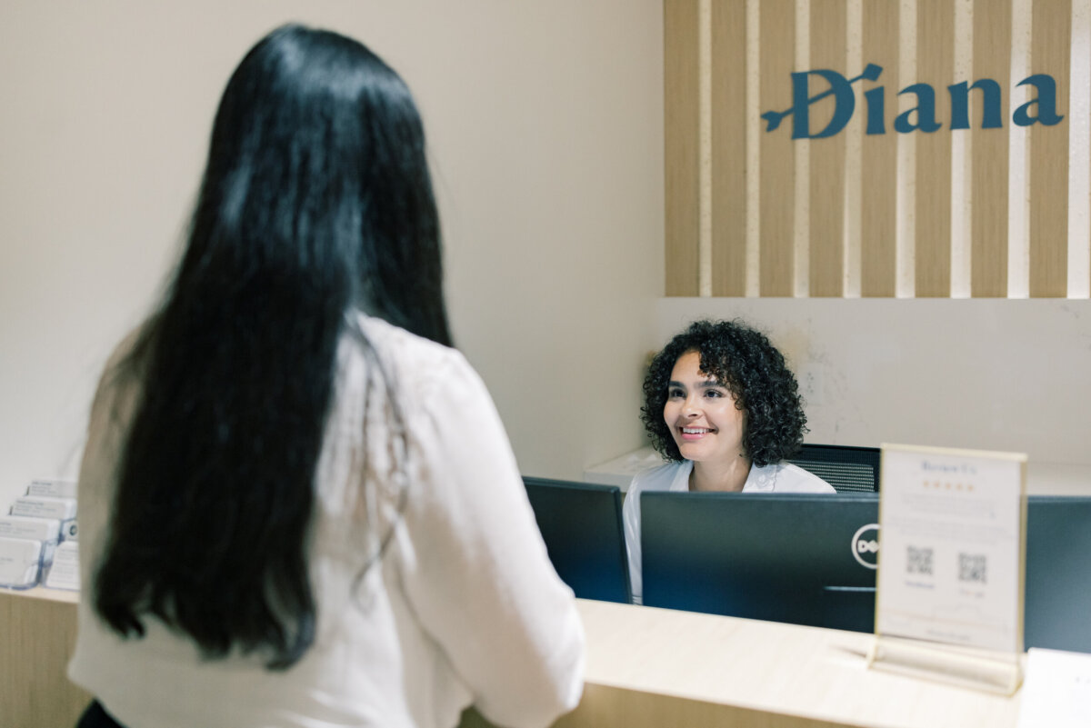 Beyond Birth: Why Women Are Choosing Midwives for Everyday Care - 5 A woman sits at a desk, greeting another woman. She is smiling and has a sign behind her that reads "Diana."