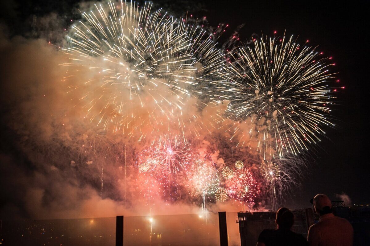 20+ Nashville Restaurants Open on July 4th - 3 Large, colorful fireworks explode in the night sky while two people watch from a distance behind a glass barrier, celebrating July 4th, 2026 after enjoying dinner at one of the many Nashville restaurants open on this festive night.