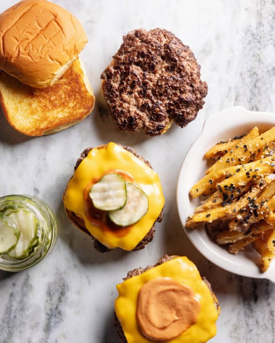 20+ Nashville Restaurants Open on July 4th - 2 An overhead view of cheeseburgers with toppings, a toasted bun, seasoned fries, and a jar of pickles on marble—perfect for a July 4th, 2026 celebration.