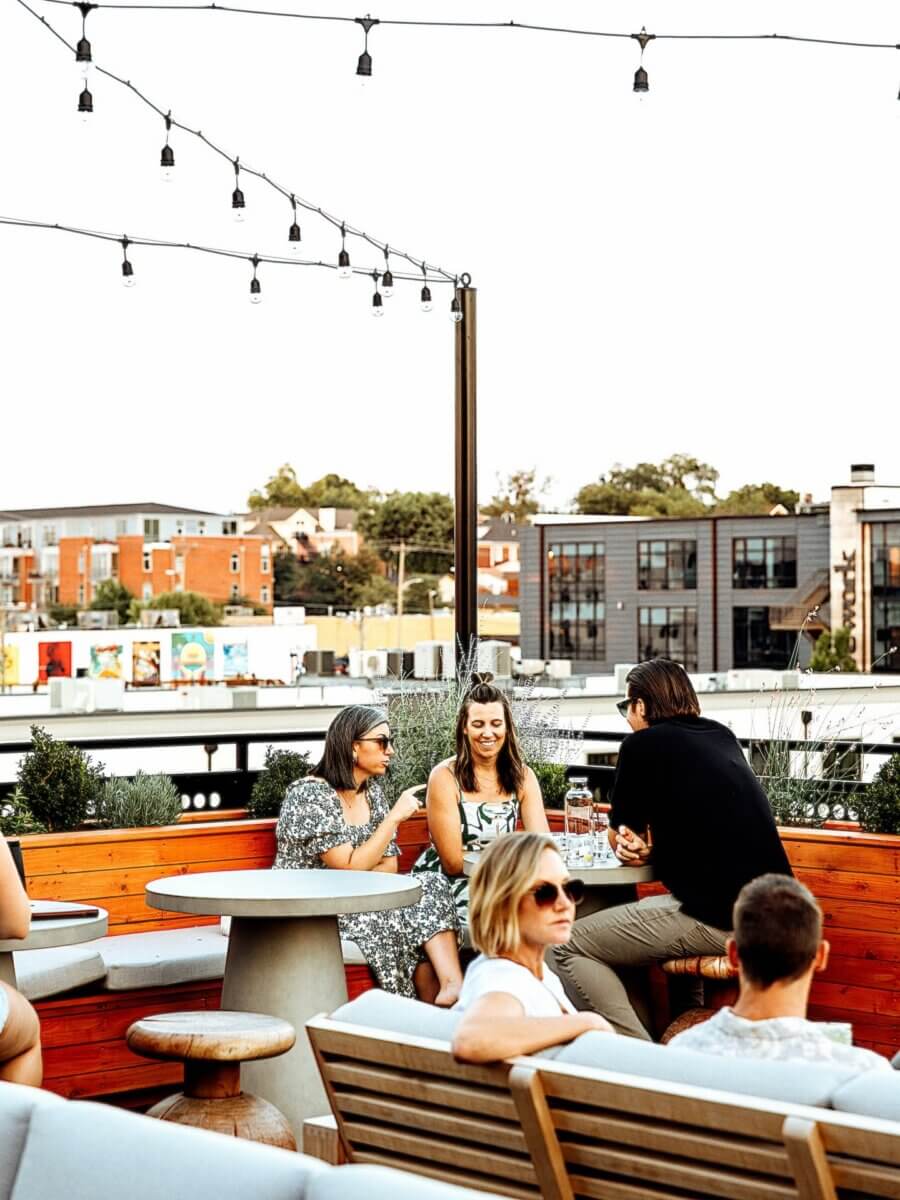20+ Nashville Restaurants Open on July 4th - 4 People sit and talk around tables on a rooftop patio with string lights, buildings, and greenery in the background—capturing the vibrant energy found at Nashville restaurants during July 4th celebrations.