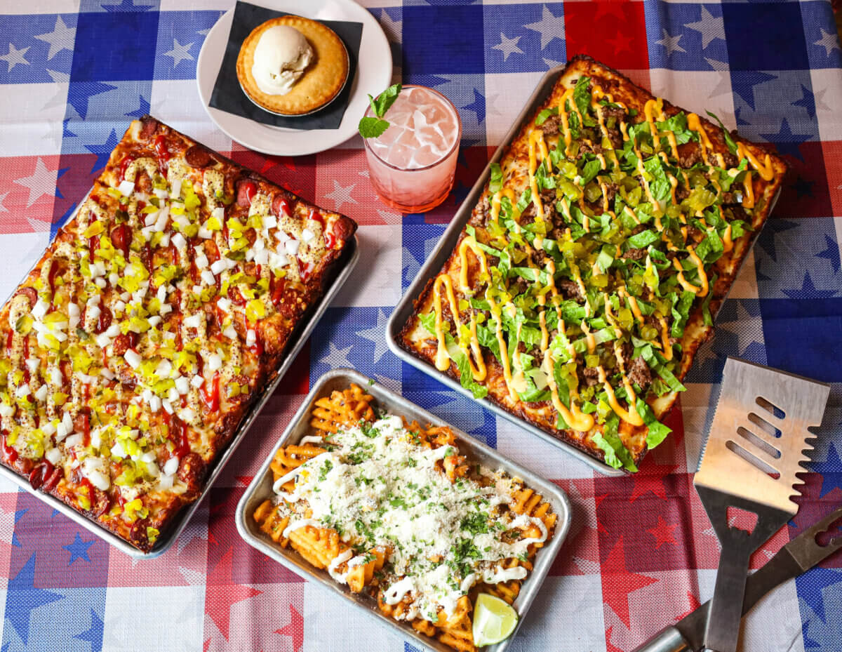 20+ Nashville Restaurants Open on July 4th - 6 Three rectangular trays of pizza with various toppings, a tray of loaded waffle fries, a pink drink with mint, and a small pie with ice cream on a July 4th-themed tablecloth, perfect for celebrating at top Nashville restaurants.