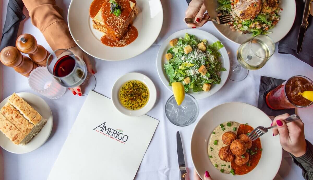 Nashville Steals, Deals & Announcements: July 2026 - 7 An overhead view of a restaurant table with Italian dishes, drinks, bread, salad, and a menu labeled "Amerigo." Diners reach for food as they enjoy Nashville deals—perfect dining for July 2026.