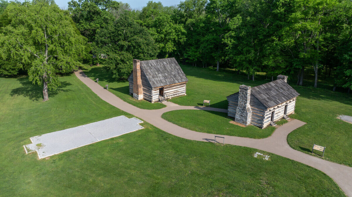 Nashville Steals, Deals & Announcements: July 2026 - 8 Aerial view of two small log cabins and a foundation outline on a grassy field, surrounded by trees and walking paths.